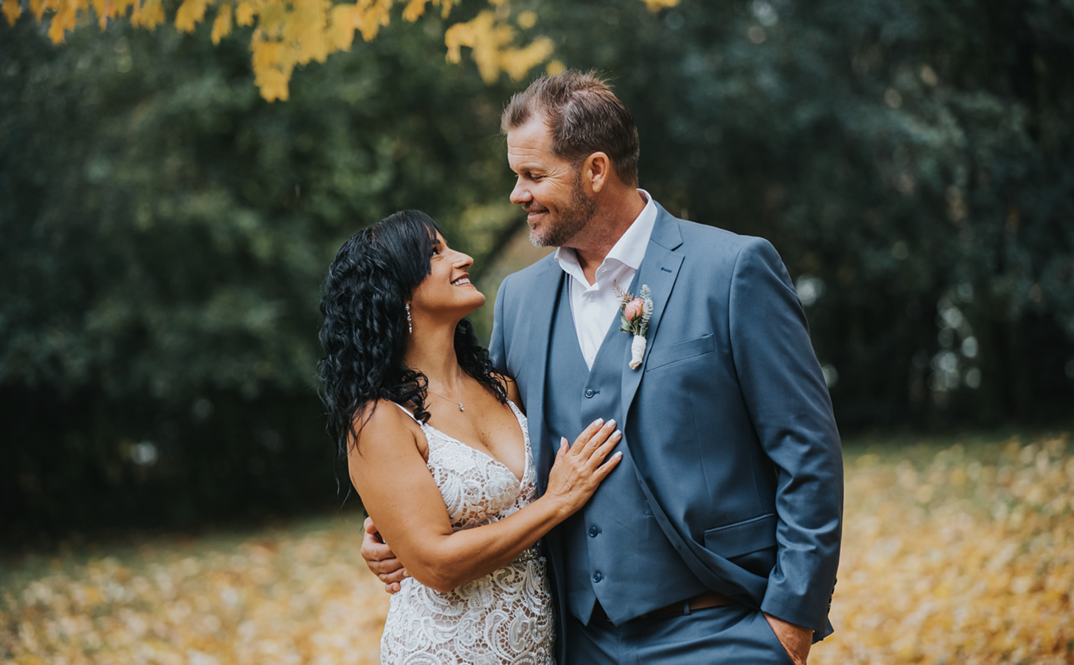 Elopement Wedding A couple stands close and smiles at each other under a tree with yellow leaves. The woman has black, curly hair and wears a sleeveless, white lace dress, while the man has short brown hair and wears a gray suit with a white shirt and a boutonniere. The background is filled with greenery. Elopements Inc